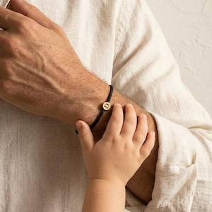 Könnte beinhalten: Ein schwarzes Kordelarmband mit einem goldfarbenen, kreisförmigen Anhänger mit dem Buchstaben "D". Das Armband befindet sich an einem Handgelenk, wobei die Hand eines Kindes ausgestreckt ist. Der Hintergrund ist hellbeige.