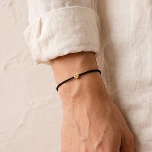 Könnte beinhalten: Ein schwarzes Kordelarmband mit einem kleinen goldfarbenen kreisförmigen Anhänger mit dem Buchstaben "M". Das Armband wird am Handgelenk getragen, mit einem hellfarbenen langärmeligen Hemd im Hintergrund.