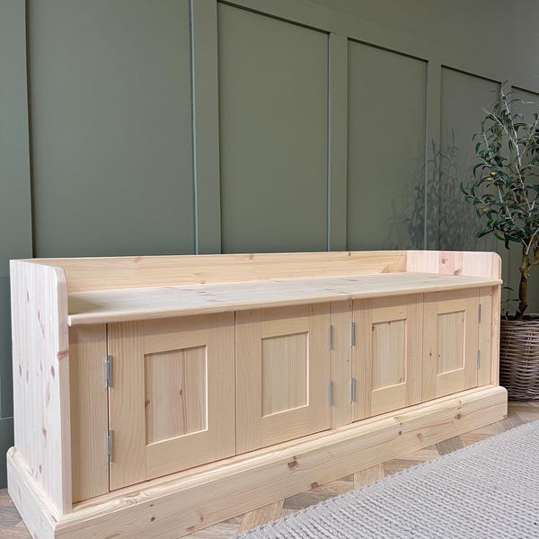 Può includere: Una panca portaoggetti in legno chiaro con quattro ante e un piano rialzato. La panca è appoggiata su un pavimento in legno con un tappeto beige chiaro di fronte.