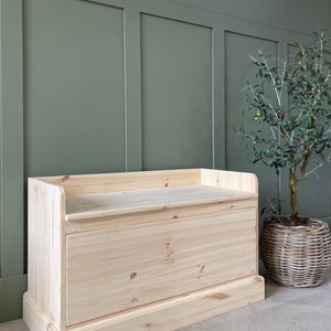 May include: A natural wood storage bench with a hinged lid and a drawer. The bench has a simple design and is made of unfinished wood.