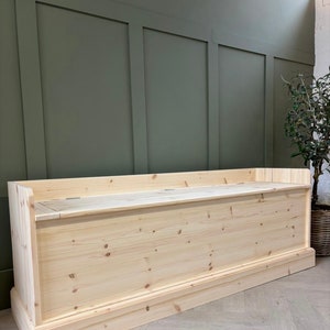 May include: A natural wood storage bench with a hinged lid. The bench is made of unfinished pine wood and has a simple, classic design.