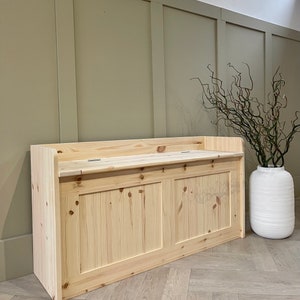 May include: A natural wood storage bench with a hinged top and two doors. The bench is made of unfinished pine wood and has a simple, rustic design.