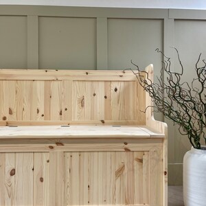 May include: A wooden bench with a hinged top and storage space. The bench is made of light-coloured wood and has a simple design. The bench is in front of a wall with green wainscoting.