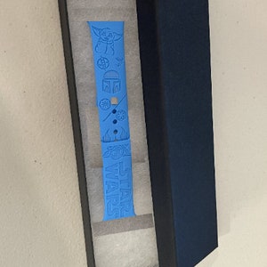 May include: A blue silicone watch band with a Star Wars theme. The band features a Baby Yoda character, a Mandalorian helmet, and the words "Star Wars". The band is in a black box with white lining.