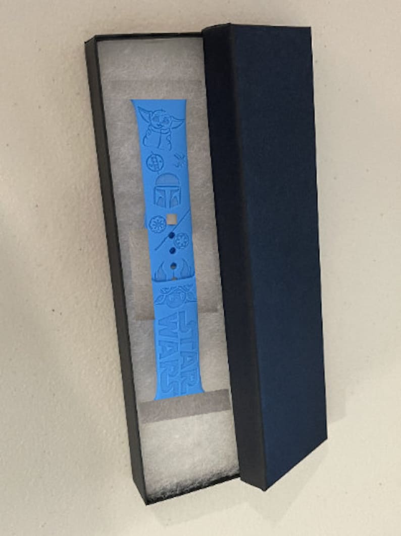 May include: A blue silicone watch band with a Star Wars theme. The band features a Baby Yoda character, a Mandalorian helmet, and the words "Star Wars". The band is in a black box with white lining.
