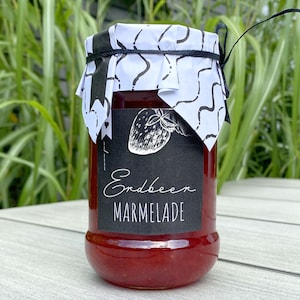 Może przedstawiać: Słoik czerwonej marmolady truskawkowej z czarną etykietą z białą ilustracją truskawki i napisem "Erdbeer Marmelade". Słoik jest zwieńczony wzorzystą biało-czarną papierową osłoną, związaną czarnym sznurkiem.
