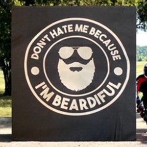 May include: A black and white graphic design on a black background with the text "Don't hate me because I'm Beardifull" in a circle with a bearded man wearing sunglasses inside the circle. A person wearing a motorcycle helmet and riding a motorcycle is standing in front of the graphic design.