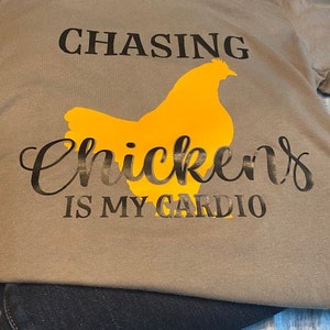 以下が含まれることがあります： 黒い文字で「Chasing Chickens is my Cardio」と書かれた茶色のTシャツ。Tシャツの中央には、黄色の鶏のシルエットがあります。