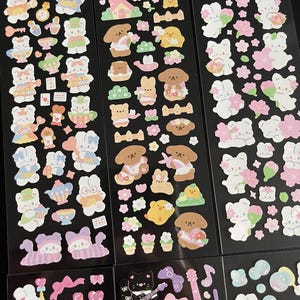 May include: A sheet of colorful stickers featuring cute cartoon characters, including bunnies, dogs, and cats. The stickers are arranged in rows on a black background. Some stickers have a floral theme, while others have a more whimsical design.