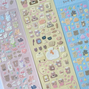 Puede incluir: Tres hojas de pegatinas con ositos de dibujos animados adorables en varias poses y actividades. Las pegatinas est&aacute;n sobre un fondo rosa claro, amarillo claro y azul claro. El texto en las pegatinas dice "무결점 곰팡" y "VOLZZZI".