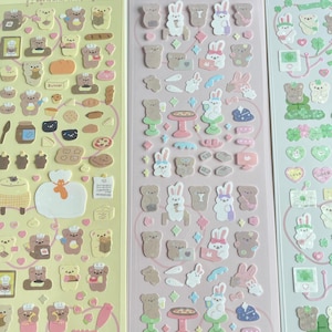 Puede incluir: Tres hojas de pegatinas con osos y conejos de dibujos animados. Las pegatinas est&aacute;n sobre un fondo rosa y amarillo. La primera hoja presenta osos horneando, la segunda hoja presenta conejos con caf&eacute; y pizza, y la tercera hoja presenta conejos con corazones y tr&eacute;boles.