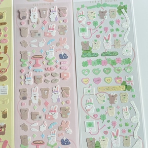 Puede incluir: Dos hojas de papel de pegatina rosa y blanco con osos y conejos de dibujos animados lindos. Las pegatinas est&aacute;n decoradas con corazones, tr&eacute;boles de cuatro hojas y otros peque&ntilde;os detalles. El texto en las hojas de pegatinas dice "Volzzi Heart Store".