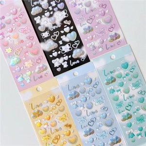 May include: Six sheets of iridescent holographic stickers featuring hearts, clouds, bows, bells, and the word "love". The stickers are in various colours, including pink, blue, yellow, and green.