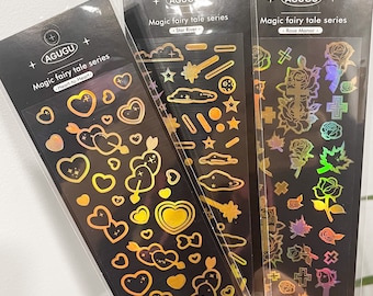 May include: Three vertical sheets of holographic stickers labeled AGUGU Magic fairy tale series. The sheets feature gold and iridescent designs including hearts with arrows, clouds, stars, wands, roses, crosses, and leaves on black backing cards.
