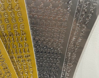 May include: A set of gold and silver alphabet stickers. The stickers are in a cursive font and are arranged in rows on a clear plastic sheet. The stickers are ideal for adding a touch of elegance to any project.
