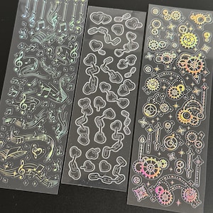 May include: Three clear sticker sheets with various designs. One sheet features musical notes and symbols, another has linked hearts, and the third showcases gears and starbursts. The designs have a holographic, iridescent finish.