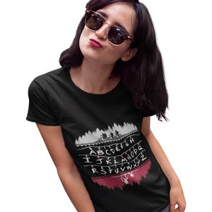 May include: Black t-shirt featuring a graphic design from the TV show Stranger Things. The design includes a forest silhouette, bicycles, string lights with the alphabet, and a red upside-down silhouette.