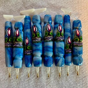 May include: A collection of eight diamond painting pens. The pens have blue and white marbled bodies with a floral design in the center. Each pen has a white tip and a gold-colored metal tip.
