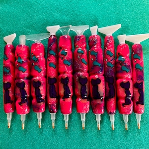 Peut inclure: Collection de dix stylos roses pour la peinture diamant, avec des pointes blanches et des pointes dorées. Les stylos présentent un motif décoratif avec des poissons turquoise et des silhouettes de chats violets. Disposés en rangée sur une surface verte.