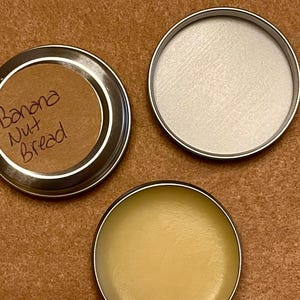 Può includere: Tre piccoli contenitori rotondi con bordi argentati su una superficie marrone. Uno è aperto, rivelando un balsamo giallo chiaro. Un altro è aperto e vuoto. Il terzo ha un'etichetta marrone con la scritta "Banana Nut Bread".