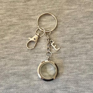Op de afbeelding: Een zilverkleurige sleutelhanger met een ronde, transparante medaillon, een karabijnhaak, een ketting en een letter "E" bedel. De medaillon is leeg, klaar om te worden gevuld met een kleine foto of aandenken. De sleutelhanger is voor sleutels of een tas.