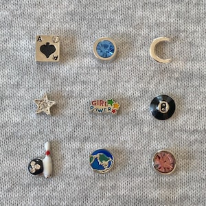 Può includere: Una collezione di ciondoli argentati con vari disegni, tra cui una stella, una mezzaluna, un birillo da bowling, un globo e una palla otto nera. Un ciondolo ha il testo "GIRL POWER". Altri ciondoli presentano una picca e gemme rotonde blu e rosa.