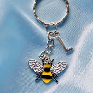 Könnte beinhalten: Ein silberner Schlüsselanhänger mit einem Bienen-Anhänger und einem Buchstaben L-Anhänger. Die Biene ist gelb und schwarz mit weißen Flügeln. Der Schlüsselanhänger befindet sich auf einem hellblauen Hintergrund.