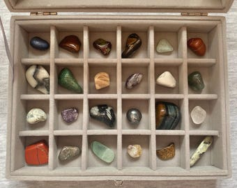Op de afbeelding: Een grijze fluwelen sieradendoos met een doorzichtig deksel, gevuld met een assortiment gepolijste edelstenen in verschillende kleuren en vormen. De edelstenen omvatten blauwe, bruine, groene en heldere stenen, gerangschikt in afzonderlijke compartimenten.
