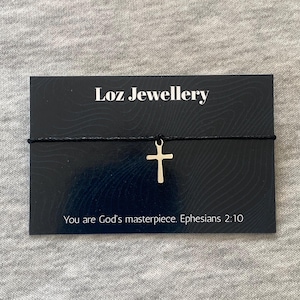 Puede incluir: Un brazalete con un colgante de cruz plateada sobre un cordón negro. El brazalete se exhibe en una tarjeta negra con el texto "Loz Jewellery" y la cita "You are God's masterpiece. Efesios 2:10."
