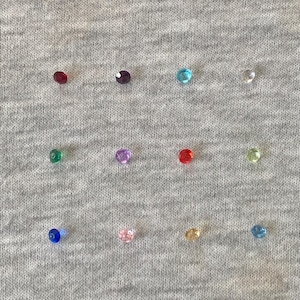 Op de afbeelding: Een verzameling kleine, kleurrijke edelstenen verspreid over een grijze stoffen ondergrond. De edelstenen zijn in verschillende kleuren, waaronder rood, paars, blauw, groen en geel. De edelstenen zijn rond en facetgeslepen en reflecteren licht.