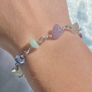 Op de afbeelding: Een handgemaakte armband met een reeks kleurrijke, gepolijste edelstenen. De stenen zijn verbonden door een zilverkleurige draad. De armband bevat stenen in tinten paars, roze, blauw en groen. De armband wordt om een pols gedragen.