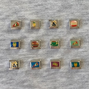 Könnte beinhalten: Eine Sammlung silberfarbener Charm-Armbänder mit farbenfrohen Emaille-Designs. Die Charms zeigen verschiedene Lebensmittel wie ein Pizzastück, einen Hamburger, eine Ananas und eine Tasse Kaffee. Weitere Charms sind ein Musikinstrument und Macarons.