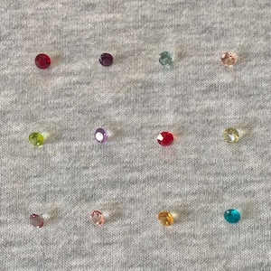 Op de afbeelding: Een verzameling kleurrijke, gefacetteerde edelstenen in verschillende kleuren, waaronder rood, paars, groen en geel. De edelstenen zijn verspreid op een grijze stoffen ondergrond, wat een sprankelend effect creëert. Deze kleine, ronde edelstenen zijn perfect voor sieraden of knutselprojecten.