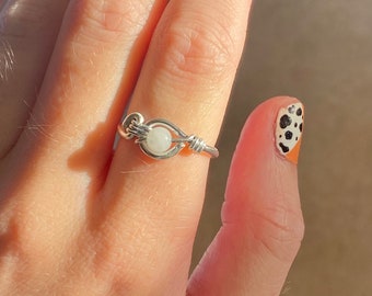 May include: A silver wire-wrapped ring with a small, round, white gemstone at the center. The ring is on a finger with a nail painted with black spots on an orange base.