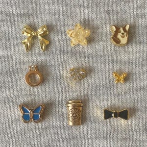 May include: A collection of small, decorative charms in various shapes, including a bow, star, ring, heart, butterfly, coffee cup, and bow tie. The charms are gold-toned with some featuring enamel details in blue and white.
