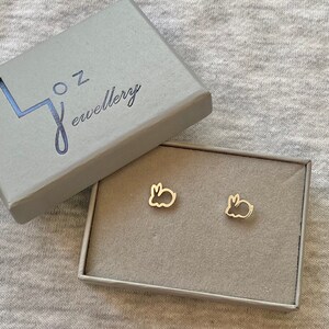 May include: Gold-colored bunny-shaped stud earrings displayed in a gray jewelry box. The box is open, revealing the earrings on a gray velvet-like insert. The box lid has the text "j oz jewellery" in a dark blue color.