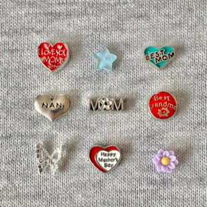 Puede incluir: Una colección de dijes decorativos de varias formas y colores. Incluye dijes en forma de corazón con textos como "I Love You Mom" y "Happy Mother's Day", una estrella, una mariposa y una flor. Los dijes son plateados, rojos, azules y morados.