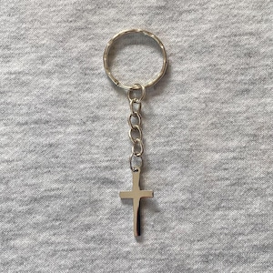 May include: A silver-colored keychain featuring a cross pendant. The cross is suspended from a chain and attached to a circular key ring. The background is a plain, light gray fabric.