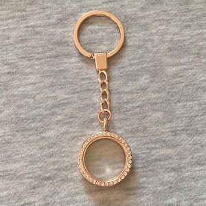 Op de afbeelding: Roségouden sleutelhanger met een ronde, transparante medaillonhanger. De hanger is omcirkeld met kleine, heldere kristallen. De sleutelhanger bevat een ketting en een ring om sleutels aan te bevestigen. De achtergrond is een grijze stof.