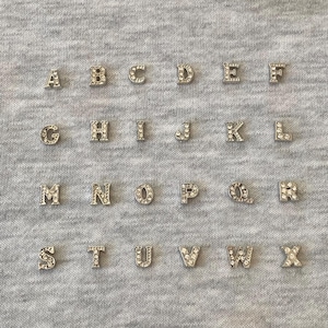 Op de afbeelding: Zilverkleurige alfabetbedels, elk bezet met kleine, heldere kristallen. De letters A t/m X zijn gerangschikt op een grijze stoffen achtergrond. Geschikt voor sieraden of knutselprojecten.