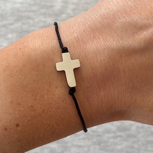 May include: A silver cross charm bracelet with a black cord. The cross is a simple, classic design, and the bracelet is worn on a wrist. The bracelet is a religious or spiritual accessory.