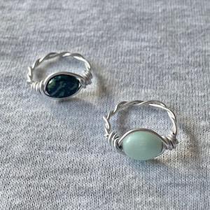 Könnte beinhalten: Zwei handgefertigte, drahtgewickelte Ringe. Ein Ring hat einen dunkelgrünen ovalen Stein, der andere einen hellgrünen ovalen Stein. Die Ringe haben ein gedrehtes Drahtband, vor grauem Hintergrund.