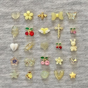May include: A selection of small decorative embellishments in various shapes, including hearts, flowers, bees, bows, butterflies, cherries, crosses, leaves, and a bunny. The embellishments are in shades of yellow, gold, clear, and red, with some featuring glitter.