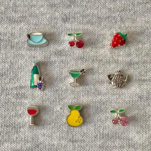 Puede incluir: Nueve pequeños dijes de esmalte de colores con bordes de metal plateado. Los dijes representan una taza de té, cerezas, una fresa, una botella de vino con uvas, un martini, una tetera, una copa de vino, una pera y cerezas.
