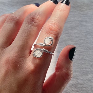 May include: A silver wire ring with two white beads. The ring is wrapped around the finger.