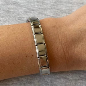 Può includere: Un braccialetto color argento con maglie rettangolari. Il braccialetto è mostrato su un polso su sfondo grigio. Le maglie sono collegate e formano una fascia flessibile.