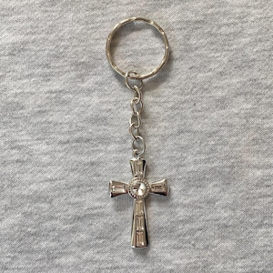 May include: Silver-colored cross-shaped keychain with a small, round, clear gem in the center. The cross has rectangular accents and is attached to a chain and a silver key ring. The background is a gray fabric.