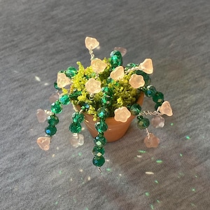 Pode incluir: Uma pequena planta em vaso com contas verdes e flores cor-de-rosa. A planta está num vaso de terracota e as contas são enfiadas em arame.