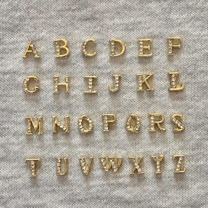 Op de afbeelding: Goudkleurige alfabetletters, elk ingelegd met kleine, heldere kristallen. De letters zijn in rijen gerangschikt op een grijze stoffen ondergrond. De letters zijn in een schreeflettertype.