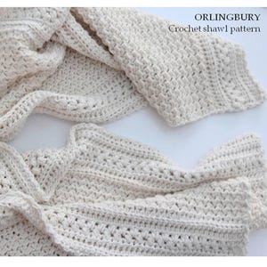 Può includere: Un modello di scialle all'uncinetto color crema. Lo scialle è realizzato con un filato morbido e strutturato e presenta un motivo a punto dettagliato. Il testo "ORLINGBURY Crochet shawl pattern" è visibile in alto a destra. Lo scialle è drappeggiato su una superficie bianca.